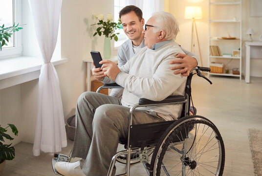 Old man being taught how to use modern smartphone by male caregiver, doesnt know how handle new technology, has problems with mobility, sitting in wheelchair. Senior people development
