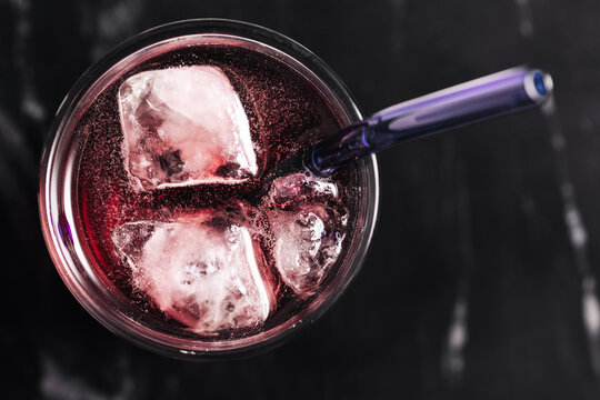 Top view of berry cocktail. Cold refreshing drink with ice cubes and glass straw. Dark marble background. Summer beverage closeup. Purple soda texture. Iced mocktail or fruit juice for hot days.