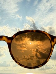 View of the sun and clouds filtered through brown-tinted sunglasses during sunset, with the sea and sky in the background.