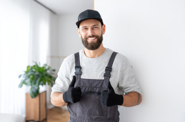 Male plumber wearing overalls and gloves stands confidently with thumbs up, ready to assist in a residential space with a welcoming atmosphere