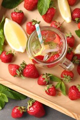 Refreshing strawberry drink with ice in mason jar, fruits and mint in wooden crate on grey table, flat lay