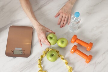 Woman at white marble table with apples, kitchen scale, dumbbells, water and measuring tape, top...
