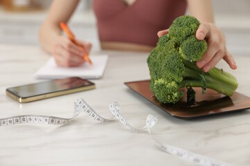 Woman developing diet plan while weighting broccoli on kitchen scale at white marble table indoors,...