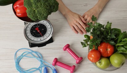 Woman at light wooden table with healthy food, kitchen scale, skipping rope and dumbbells, top view