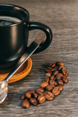 Tasse noire remplie de café sur une sous-tasse en bois avec des grains de café et une petite cuillère, le tout sur une table grise façon bois