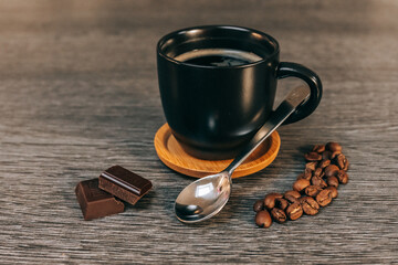 Tasse noire remplie de café sur une sous-tasse en bois avec des grains de café, une petite cuillère et des carrés de chocolat, le tout sur une table grise façon bois