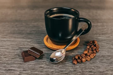 Fototapeten Tasse noire remplie de café sur une sous-tasse en bois avec des grains de café, une petite cuillère et des carrés de chocolat, le tout sur une table grise façon bois © Damien