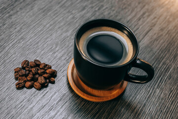 Tasse noire remplie de café sur une sous-tasse en bois avec des grains de café, le tout sur une table grise façon bois