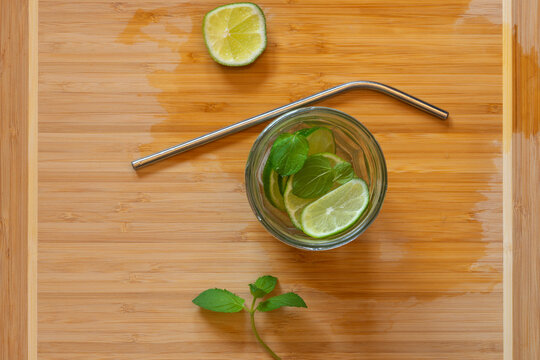 Mojito-like refreshing lemonade with mint, lime and ice in a jar with a metal straw on wet wooden cutting board against contrast background, top view