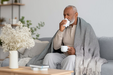 a Black man sneezing with a tissue, at home sitting on his couch in his living room wearing pajamas and a blanket over his shoulders and coffee cup in his hand, books on the table in front of him.