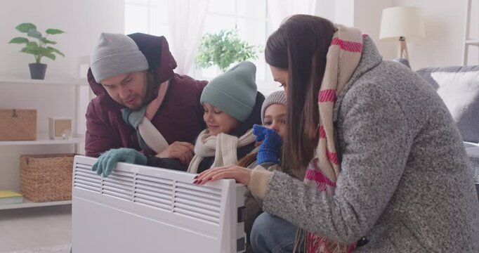 Freezing parents and children wearing warm clothes sitting near electric heater and trying to warm up. Young family with heating problems suffering from low temperature, calling emergency services.