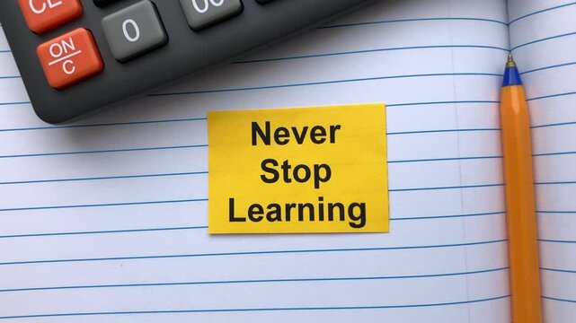 A yellow paper note with the words Never Stop Learning on it lying in an open notebook near a calculator and ballpoint pen.