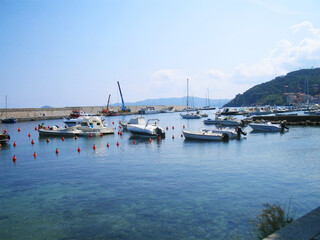A serene and picturesque view of boats gently floating in the calm waters of the port on Elba Island, Italy. Surrounded by pastel-colored buildings and rugged coastal hills, the tranquil harbor scene