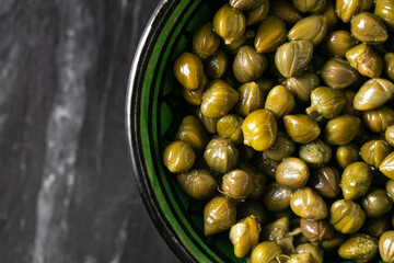 Pickled capers in ceramic bowl on dark background. Salty green buds with wrinkled skin texture....