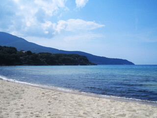 Stunning beach view on Elba Island, Italy, showcasing crystal-clear turquoise waters, golden sand, and lush Mediterranean vegetation. The peaceful shoreline, framed by gentle waves and rocky outcrops