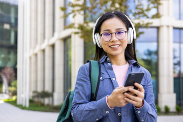 A smiling Asian woman with glasses and headphones holding a phone outdoors near a modern building.