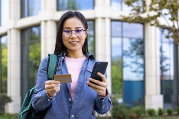 An Asian woman stands outside, holding a credit card and smartphone with a smile.