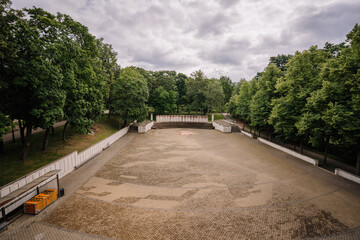 Empty open-air amphitheater with patterned stone flooring surrounded by green trees in a city park under a cloudy sky. Urban tranquility and copy space.