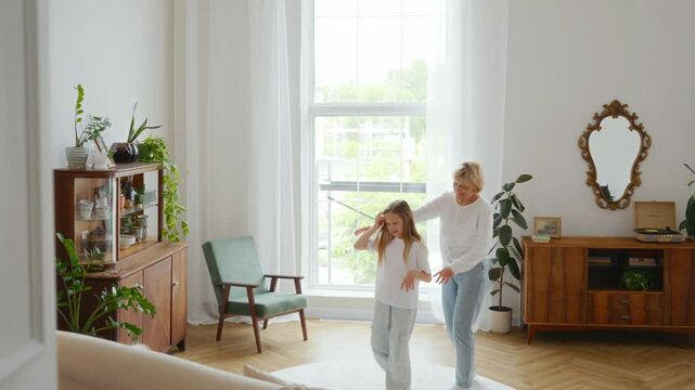 Shot through a doorway, a grandmother and granddaughter joyfully dance in a sunlit room. The frame captures warmth, activity, and intergenerational bonding beyond the screen. High quality 4k footage