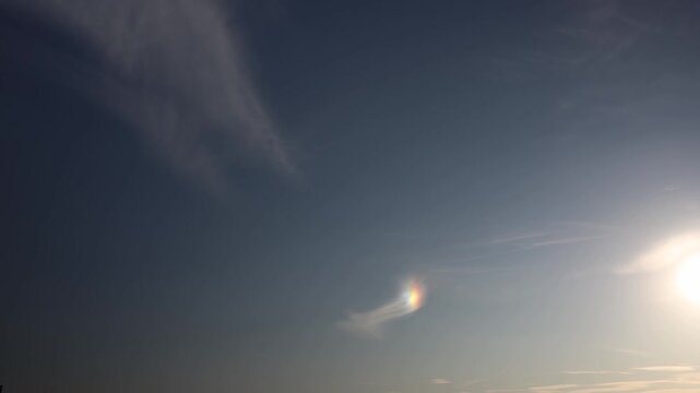 Bright sun in clear sky with rare rainbow-colored sundog glowing beside wispy clouds in peaceful early evening atmosphere
