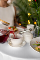 Close-up of tea being poured into a white cup on a cozy breakfast table with food, sugar bowl, and flowers. Morning dining atmosphere
