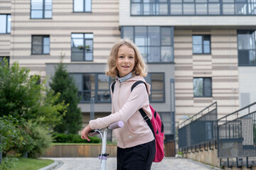 Obraz premium A cheerful girl rides her scooter towards school, wearing a backpack. She is in a modern urban setting, surrounded by green trees and buildings on a sunny day.