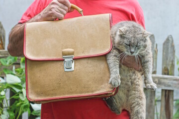 one man in red clothes holds in hand gray cat and brown leather old briefcase on summer street