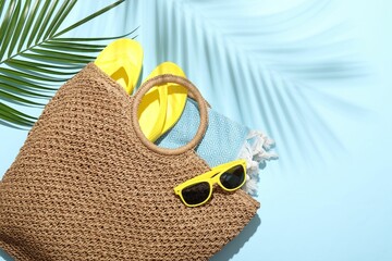 Wicker bag with beach towel, sunglasses, flip flops and palm leaf on light blue background, flat...