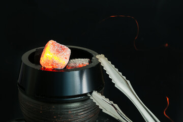 Smoldering charcoal cubes in hookah holder and tongs on black background, closeup