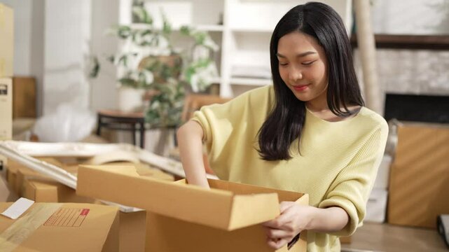 Young entrepreneur packing product in cardboard box at home office with package scattering around. Attractive online seller carrying package and preparing for delivery and shipping order. Myrmidon.