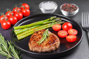 Tasty grilled beef steak served with green asparagus and cherry tomatoes on black plate, with cutlery. Perfect for food menu, diet, or culinary design concepts.