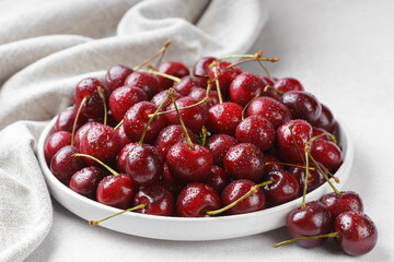 Close up of white plate filled with ripe red sweet cherry covered in fresh water drops, placed on light linen napkin on white background. Delicious food, juicy summer fruit and healthy eating.