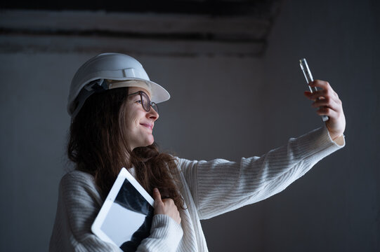 Female architect wearing a safety helmet taking a selfie with her smartphone during a site inspection while holding a tablet