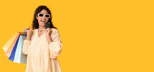 Happy young African-American woman with shopping bags and credit card on yellow background with space for text