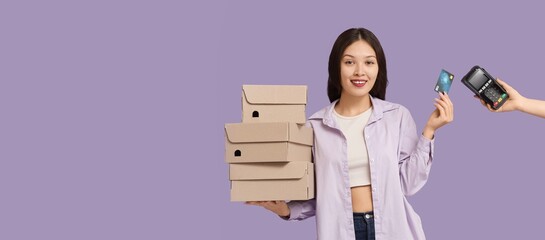Young Asian woman with boxes and credit card paying via terminal on lilac background with space for...