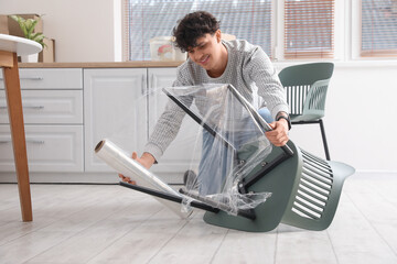 Young man wrapping chair with stretch film in kitchen