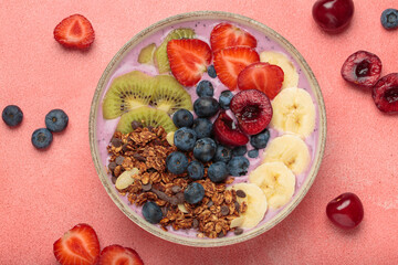 Vibrant and nutritious acai bowl topped with sliced kiwi, strawberries, blueberries, cherries, banana, and granola, a colorful and healthy meal or breakfast on a pink background