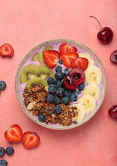 Vibrant and nutritious acai bowl topped with sliced kiwi, strawberries, blueberries, cherries, banana, and granola, a colorful and healthy meal or breakfast on a pink background