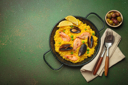 Seafood paella with rice, shrimp, mussels, peas, and lemon wedges served in a black traditional pan on a green rustic background with olives and cutlery, copy space