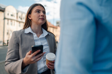 Businesswoman using smartphone and holding coffee, talking to colleague outdoors