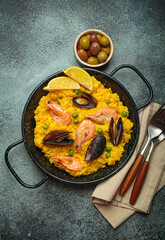 Seafood paella with rice, shrimp, mussels, peas, and lemon wedges served in a black traditional pan on a dark rustic background with olives and cutlery
