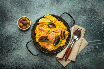 Seafood paella with rice, shrimp, mussels, peas, and lemon wedges served in a black traditional pan on a dark rustic background with olives and cutlery