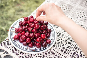 A hand reaches for fresh, ripe cherries on a white plate, ready to be enjoyed. A hand reaching for ripe cherries on a plate atop a delicate lace tablecloth, captured in natural light.