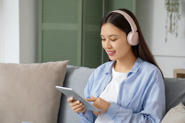 Beautiful young Asian woman in headphones listening to audiobook with modern tablet computer and...