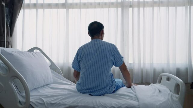 Rear view of sick old man sitting on bed looking at window inside hospital in lonely, depressed and hopeless attitude, health concept.