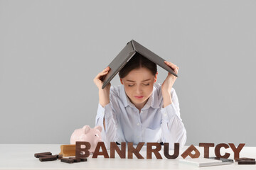 Stressed businesswoman with laptop and word BANKRUPTCY at table on light background