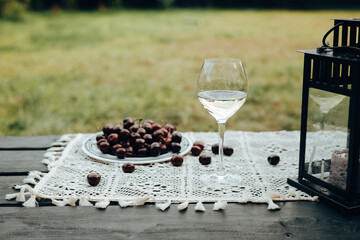 White wine in a glass with fresh cherries on a lace tablecloth outdoors in the summer. A plate of cherries on a lace tablecloth, alongside a decorative lantern on a wooden table with grass background.