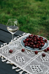 A glass of white wine and a plate of cherries on a table outside on a sunny day. A refreshing glass of white wine alongside a plate of ripe cherries on a table outdoors, ready for enjoyment.