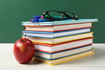 Different school stationery, glasses and red apple on white wooden table against blackboard, closeup