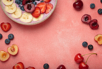 Vibrant arrangement of fresh fruit including strawberries, cherries, blueberries, and bananas surrounds a yogurt bowl on a textured pink surface with copy space